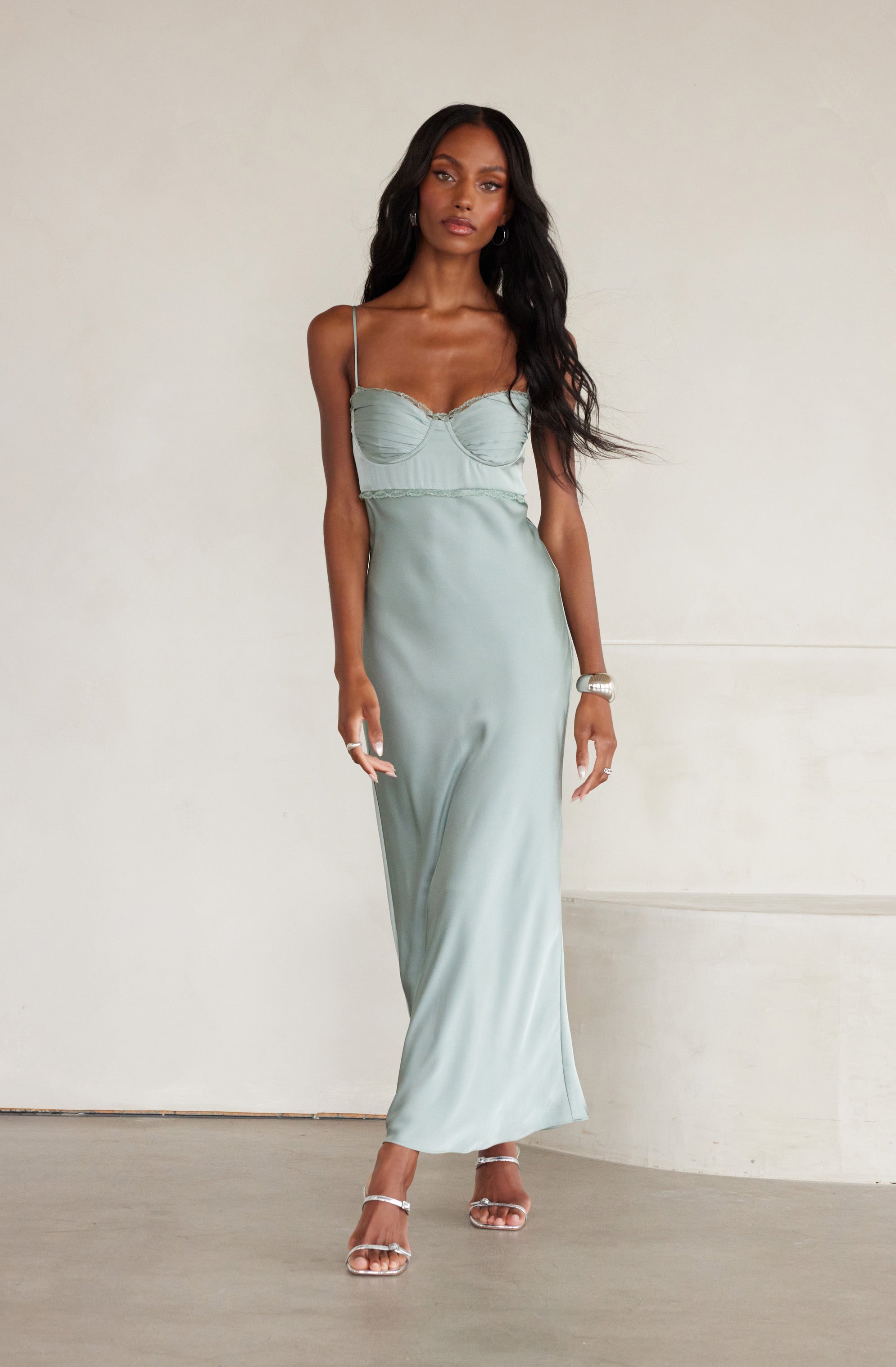 The woman is wearing a light green satin maxi dress with thin spaghetti straps. The dress features a fitted bodice with ruching detail and a slightly gathered waist. She is also wearing clear high-heeled sandals and accessorizing with a bracelet on her right wrist and earrings.