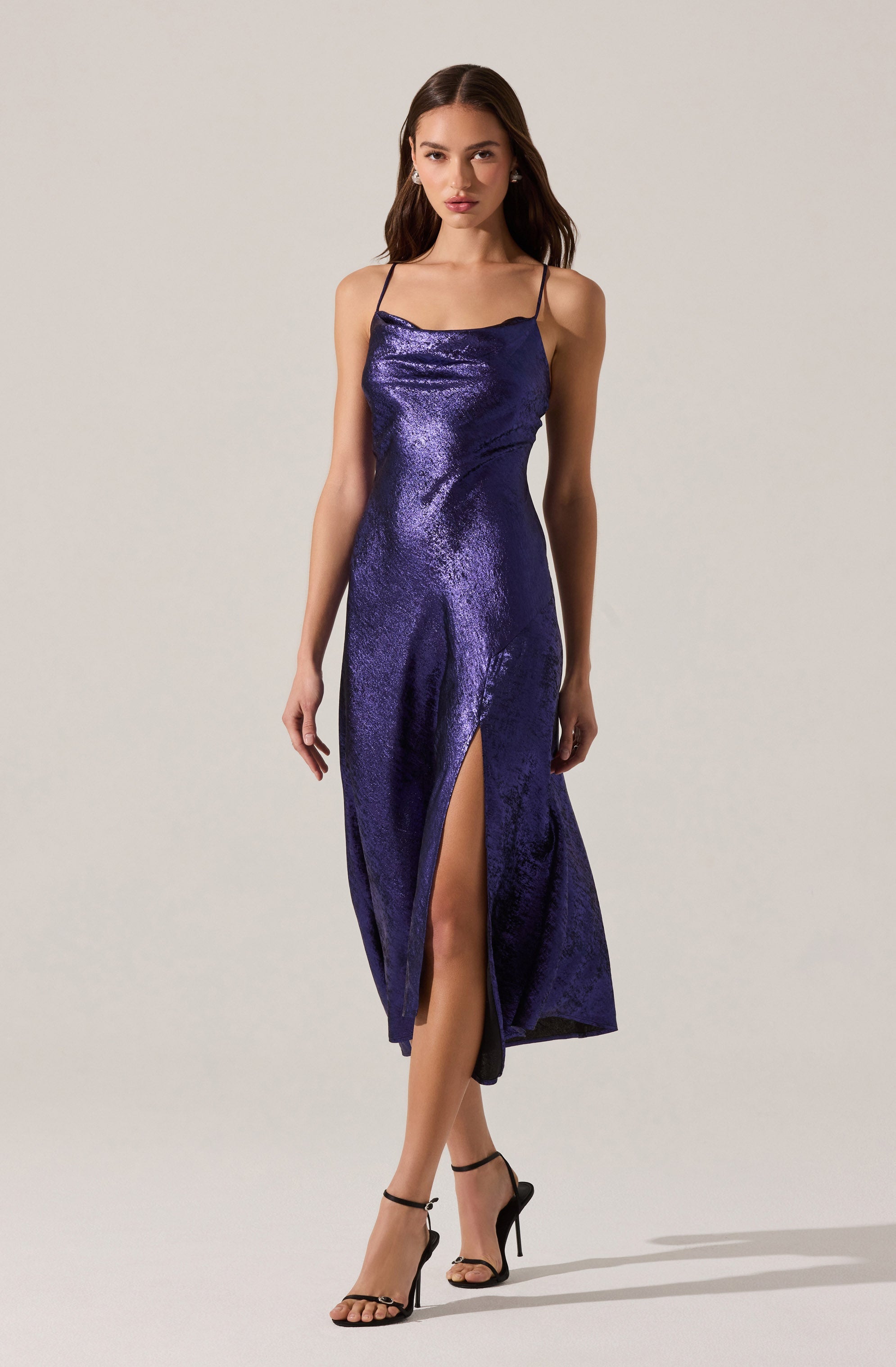 A woman is wearing a shiny, deep purple dress with a satin-like finish. The dress has a fitted bodice with spaghetti straps and a straight neckline. It features a high slit on the front left side, revealing her leg, and falls to a midi length. She is wearing black high-heeled sandals with ankle straps.