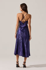 A woman is wearing a shiny, dark purple satin midi dress with thin spaghetti straps. The back of the dress features intricate criss-cross and tie details, creating an open-back design. She has on black high-heeled sandals with ankle straps.