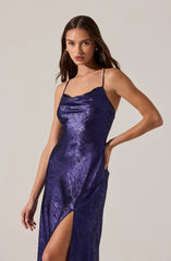 A woman is wearing a shiny, metallic purple slip dress with thin spaghetti straps. The dress features a subtle cowl neckline and a high slit on one side, revealing her leg. The fabric has a textured, crushed velvet appearance, contributing to its shimmering effect.