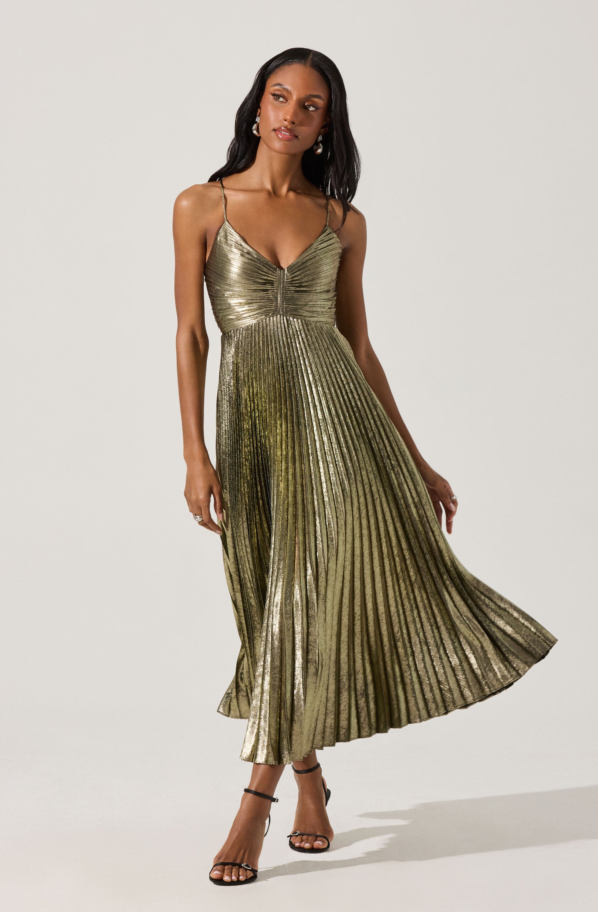 A woman is wearing a metallic gold pleated dress with thin spaghetti straps. The dress features a fitted bodice with a slight V-neckline and a flowing A-line skirt that reaches her ankles. She is also wearing black strappy high-heeled sandals.