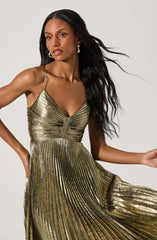 A woman is wearing a metallic gold pleated dress with thin spaghetti straps. The dress has a fitted bodice with a gathered detail at the center and flows into a full length skirt with vertical pleats. She accessorizes with large, dangling earrings and has long, wavy black hair.