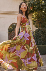 A woman is wearing a sleeveless, V-neck, ankle-length dress with a vibrant, nature-inspired floral pattern. The dress features a mix of earthy green, purple, red, and beige colors with pleated fabric that adds texture and movement. The pattern includes large, abstract flowers and leaves that create a bold, artistic look.