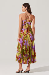 A woman is wearing a sleeveless maxi dress with a colorful, abstract floral pattern in shades of purple, green, yellow, and rust. The dress features a fitted top with thin spaghetti straps that crisscross and tie at the back, creating open sides with cut-out details. The pleated skirt flows down to her ankles, showcasing a vibrant and eye-catching design. She pairs the dress with black strappy heels.