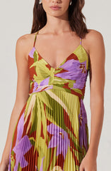 A woman is wearing a sleeveless, V-neck dress with thin spaghetti straps. The dress features a vibrant, abstract floral pattern in shades of purple, green, red, and yellow, with pleated fabric throughout.