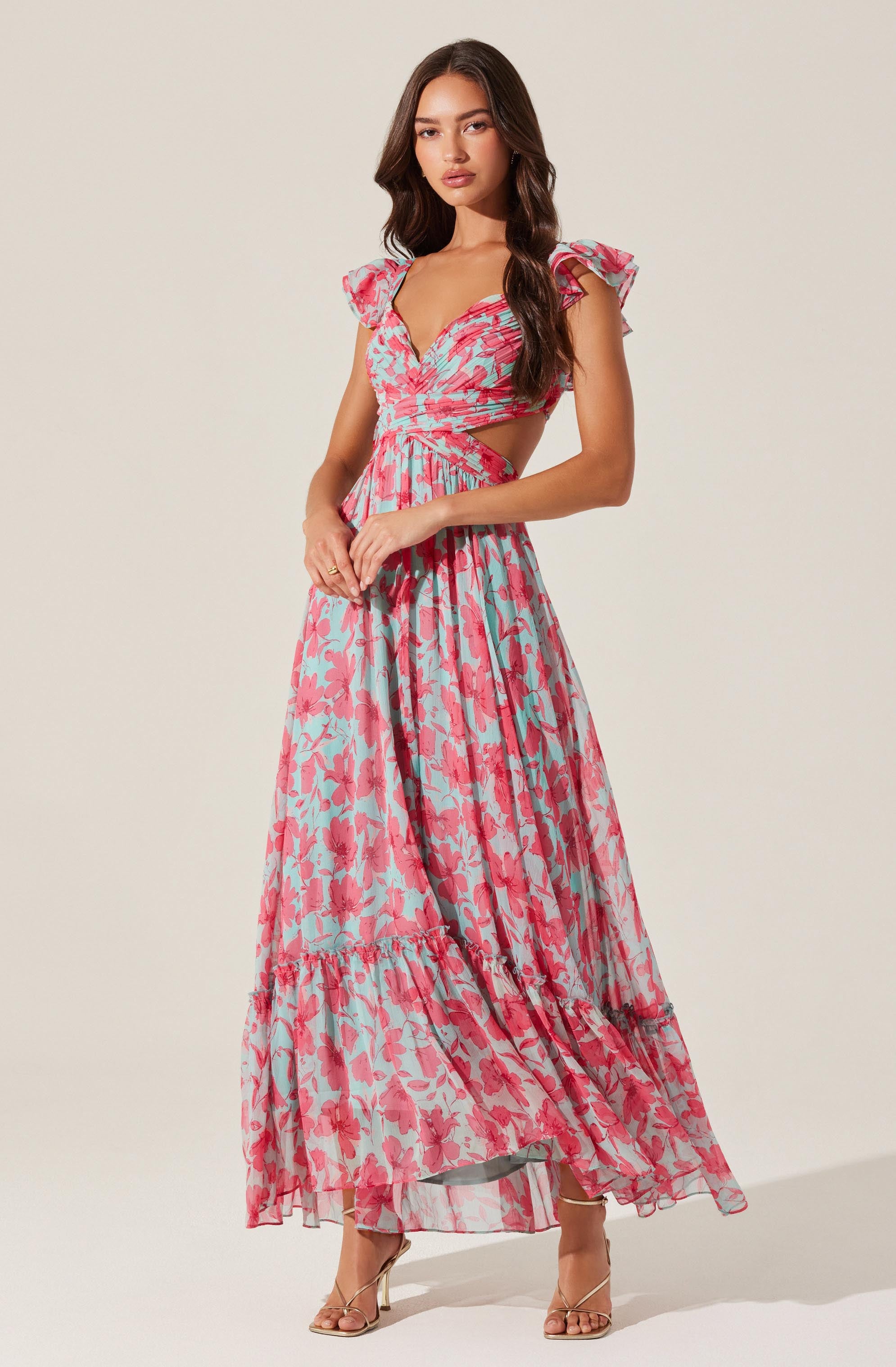 A woman is wearing a long, colorful dress with a pink, red, and light blue floral pattern. The dress features short, ruffled sleeves, a sweetheart neckline, cutouts at the waist, and a flowing, tiered skirt. She is also wearing strappy, high-heeled sandals.