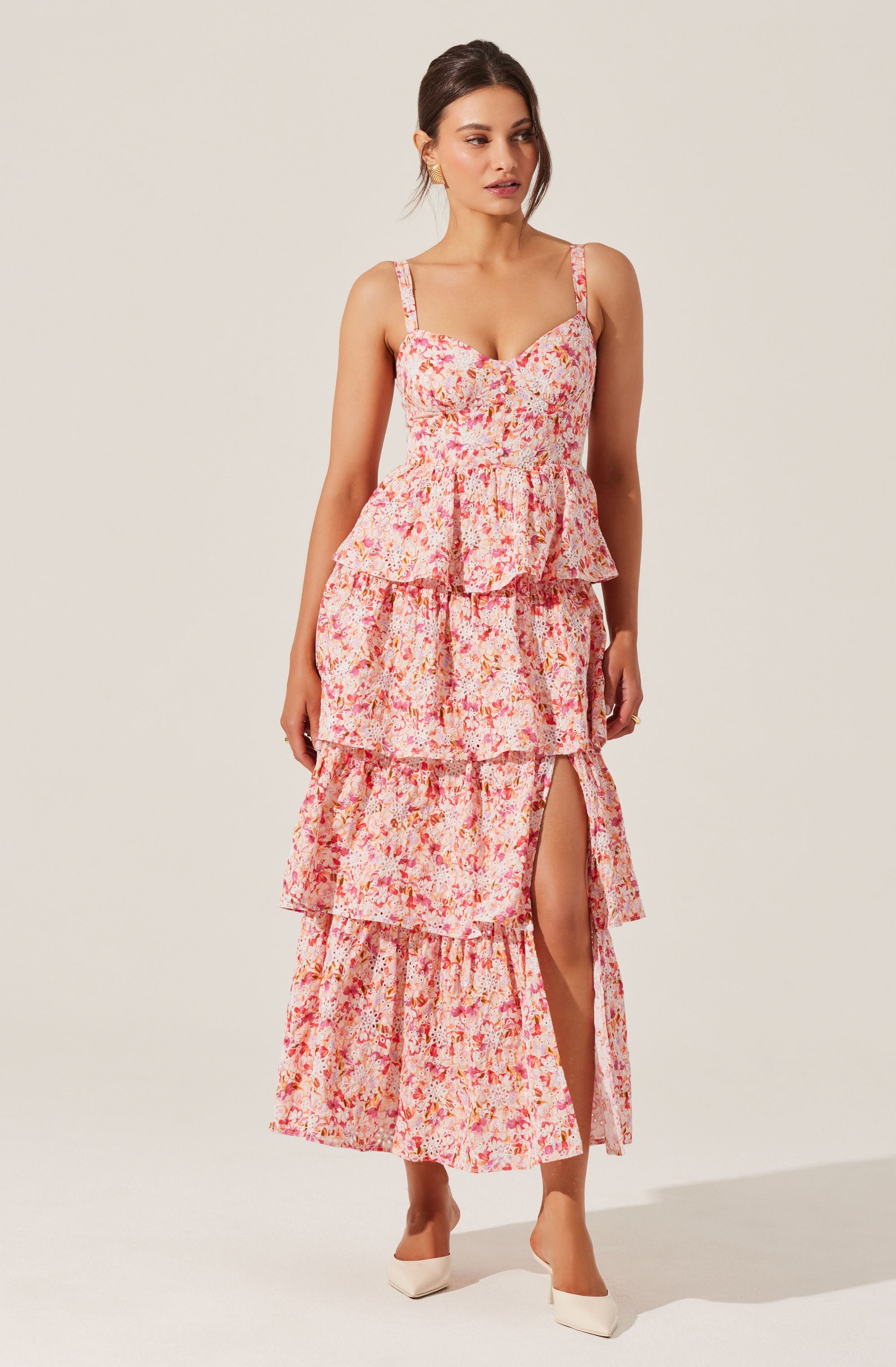 A woman is wearing a sleeveless, floral maxi dress with a pink, orange, and white pattern. The dress has a fitted bodice with thin straps and a tiered, ruffled skirt that features a front slit revealing her leg. She is also wearing white pointed-toe high heels.
