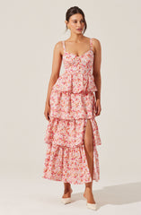 A woman is wearing a sleeveless, floral maxi dress with a pink, orange, and white pattern. The dress has a fitted bodice with thin straps and a tiered, ruffled skirt that features a front slit revealing her leg. She is also wearing white pointed-toe high heels.