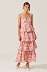 A woman is wearing a sleeveless, floral print maxi dress with multiple ruffled tiers throughout the skirt. The dress features a fitted bodice with a sweetheart neckline and thin shoulder straps. She pairs the dress with white pointy-toed high heels and gold earrings.