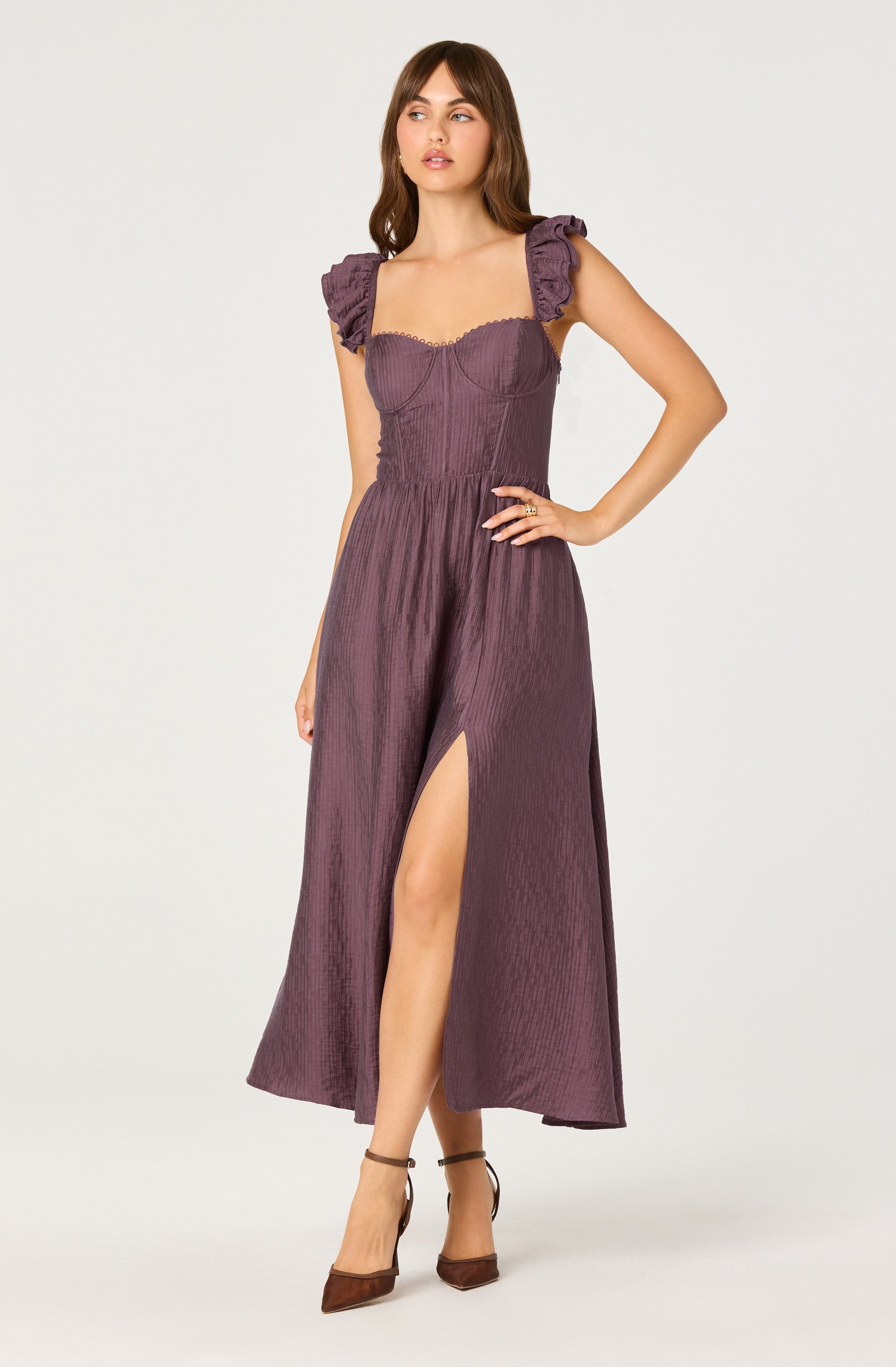 A woman is wearing a long, purple dress with textured fabric. The dress features a fitted bodice with sweetheart neckline and ruffled shoulder straps. The skirt is flowing with a high slit on the left side. She is wearing brown high-heeled shoes.