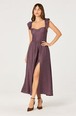 A woman is wearing a long, purple dress with textured fabric. The dress features a fitted bodice with sweetheart neckline and ruffled shoulder straps. The skirt is flowing with a high slit on the left side. She is wearing brown high-heeled shoes.