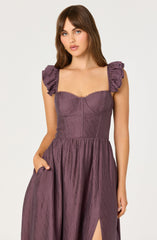 A woman is wearing a mauve-colored dress with short, ruffled sleeves and a fitted bodice featuring stitched detailing. The skirt is full and gathered, with a slit on one side. The fabric has a textured, slightly shiny appearance.