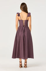 The dress is a mauve-colored, floor-length gown made of a shiny, textured fabric. It features ruffled shoulder straps and a fitted bodice with a lace-up back detail secured with small metal eyelets and a matching fabric tie. The skirt flows loosely to the ground, and the model is wearing strappy high-heeled sandals.