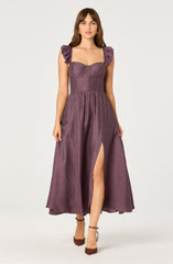 A woman is wearing a long purple dress with a fitted bodice and ruffled shoulder straps. The dress has a textured, shiny fabric, features a slit front revealing her leg, and has a slightly gathered waistline. She is also wearing brown ankle-strap heels.
