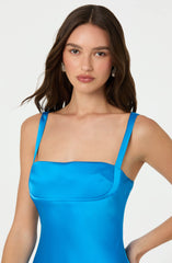 A woman is wearing a bright blue satin dress with thin spaghetti straps. The dress has a smooth, shiny texture and a structured bust with a slight gathering at the top. She has shoulder-length, wavy brown hair and is accessorized with pearl earrings.