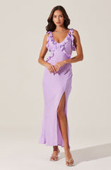 A woman is wearing a sleeveless, lavender-colored maxi dress with ruffled straps and a V-neckline. The dress features a thigh-high slit on one side, revealing her leg. She is also wearing open-toe heels with a transparent strap and a necklace.