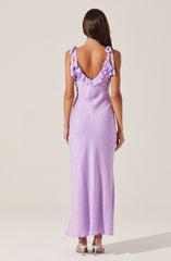 A woman is wearing a lavender maxi dress with a textured fabric. The dress features ruffled shoulder straps tied into bows and a deep V-back. She has long, straight brown hair and is accessorized with a necklace. She is also wearing high-heeled shoes.