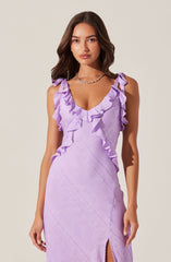 A woman is wearing a sleeveless, lavender-colored dress with ruffle detailing along the neckline and shoulders. The dress has a fitted silhouette, a V-shaped neckline, and a small slit on the front. The fabric appears to have a textured, subtle pattern. She is accessorized with a silver chain necklace.