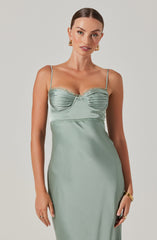 A woman is wearing a light green satin slip dress with thin spaghetti straps. The bodice features ruched detailing with a lace trim along the top edge. The dress is fitted and smooth, extending down without visible embellishments. She accessorizes with gold hoop earrings, a gold bracelet, and rings.