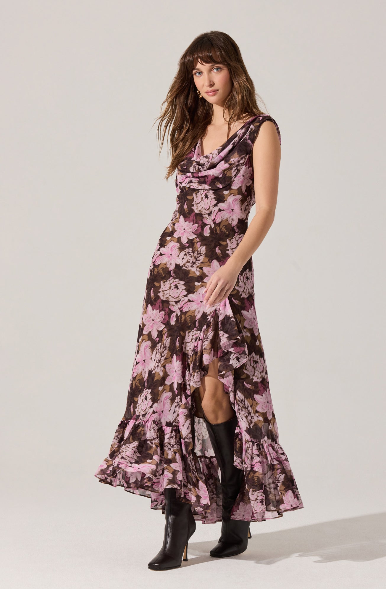 A woman is wearing a sleeveless, ankle-length dress with a floral pattern consisting of pink and purple flowers on a dark background. The dress features a draped neckline and ruffled hem at the bottom. She is also wearing black high-heeled boots.