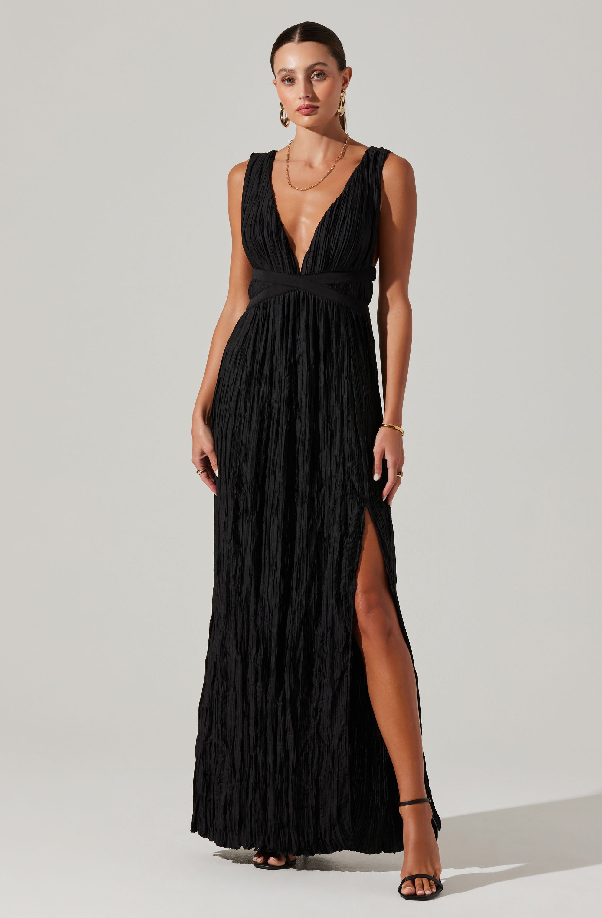 A woman is wearing a long, black dress with a deep V-neckline and wide shoulder straps. The fabric of the dress is textured with a crinkled appearance. The dress features a high slit on the left side, revealing her leg and black high-heeled sandals.