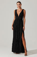 A woman is wearing a long, black dress with a deep V-neckline and wide shoulder straps. The fabric of the dress is textured with a crinkled appearance. The dress features a high slit on the left side, revealing her leg and black high-heeled sandals.