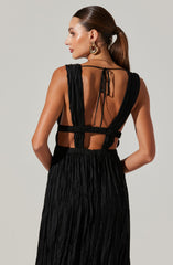 The image shows a woman wearing a sleeveless black dress with a sheer, open-back design. The upper part of the dress features wide, gathered straps that extend over her shoulders and connect at the back with a thin tie. The waistline is defined, and the skirt is made of a textured, crinkled fabric that flows down. She accessorizes with large, gold hoop earrings and has her hair pulled back into a sleek ponytail.