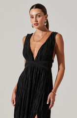 A woman is wearing a sleeveless black dress with a deep V neckline and a gathered, pleated fabric texture. The dress has a wide waistband that accentuates her waist and a subtle slit at the bottom. She is accessorized with gold hoop earrings, a gold chain necklace, and a gold bracelet.