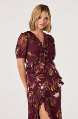 The dress is made of a silky fabric with a dark burgundy base color featuring a floral pattern in shades of yellow, pink, white, and green. It has short puffed sleeves with elastic cuffs, a V-neckline, and a ruffled front with a tie detail at the neck. The skirt has a high slit with ruffles along the edges.