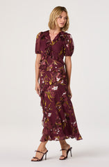 A woman is wearing a long, burgundy dress with a floral pattern featuring white, yellow, and pink flowers and green leaves. The dress has puffed short sleeves, a V-neckline, and a ruffled, layered skirt with a ruffle around the waist. She is also wearing strappy high-heeled sandals.