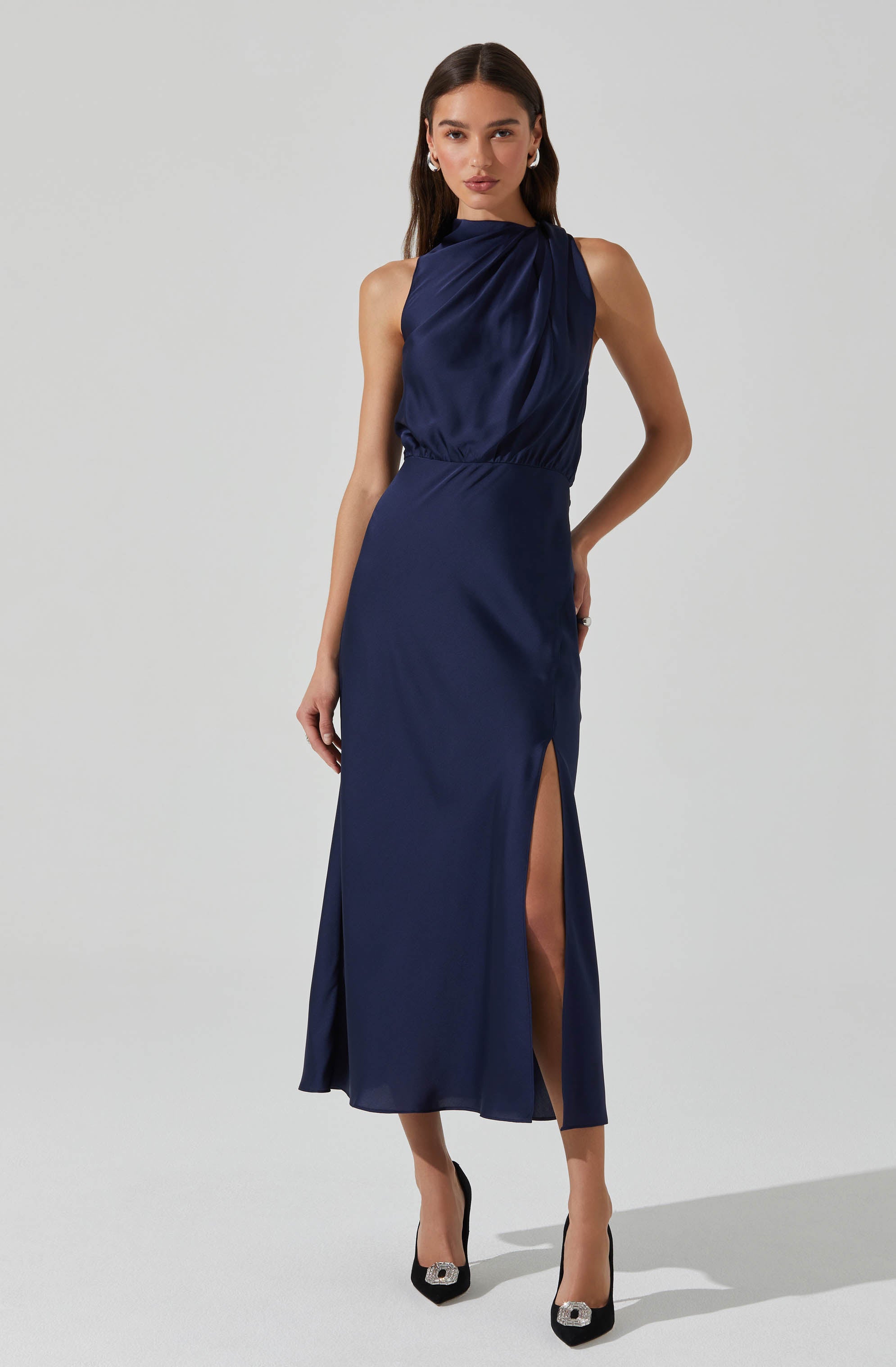 A woman is wearing a sleeveless, high-neck, navy blue satin dress. The dress has a gathered, draped bodice and a fitted waist. The skirt is long and features a side slit that reveals one leg. She is also wearing black high heels with decorative embellishments on the toes.