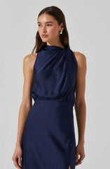 A woman is wearing a sleeveless, navy blue satin dress with a high neckline. The top part of the dress has gathered fabric that drapes diagonally across her chest, creating a soft, flowing look. The waistline is fitted, and the skirt is smooth with a slight slit on one side. She has her hair styled down and is wearing silver earrings.