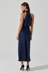 A woman is wearing a sleeveless, navy blue, satin-like gown with a high neckline that drapes around her neck. The dress has a fitted waist and flows down to her ankles. She is also wearing black high-heeled shoes with beige soles.
