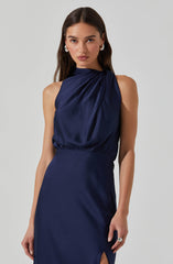A woman is wearing a sleeveless, dark navy blue satin dress with a draped, gathered neckline that creates soft folds across the chest. The dress is fitted at the waist and has a small slit on the side. She is accessorized with large, hoop earrings and a ring on her right hand.