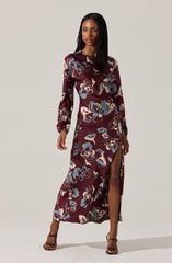 A woman is wearing a long, floral dress with a high slit. The dress has a maroon background with large, abstract floral patterns in shades of blue, beige, and tan. It features long sleeves with gathered cuffs and a round neckline.