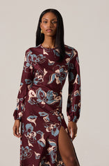 A woman is wearing a long-sleeved, floral-print dress with a high neckline. The dress features a dark maroon base color with large, abstract flowers in shades of blue, beige, and cream. It has a high slit on one side, revealing her leg. The sleeves are gathered at the wrists, creating a slight puffed effect.