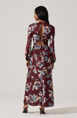 A woman is wearing a long, floral-printed dress with a burgundy background and large blue and cream flowers. The dress features long, puffed sleeves and a back slit. The back of the dress has a distinctive open design with a tie-up detail at the top and a lace-up closure in the middle.