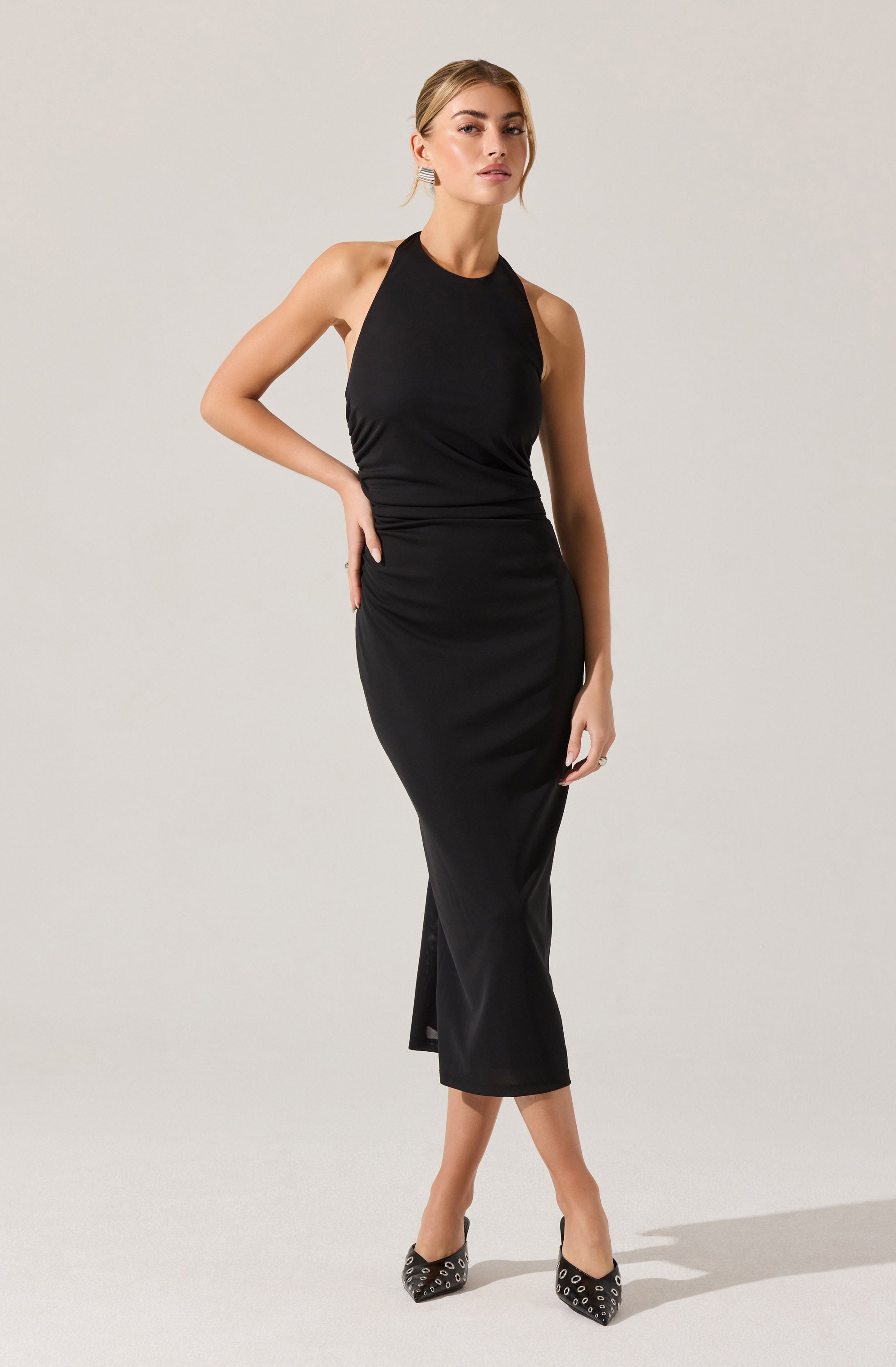 A woman is wearing a long, black, sleeveless dress with a high neckline. The dress is fitted through the waist and hips, elongating the figure, and has a slight slit at the front hem. She is also wearing black pointed heels with silver eyelet details.