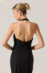 A woman is wearing a sleeveless, backless black evening gown with a halter neckline. The fabric of the dress is smooth and slightly shiny, gathered at the waist with draping detail. She has styled her hair in an elegant updo and is wearing small, stud earrings.