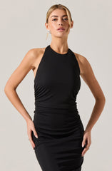 A woman is wearing a sleeveless, high-neck black dress that is fitted and ruched around the waist and hips. She is also wearing large, rectangular silver earrings.