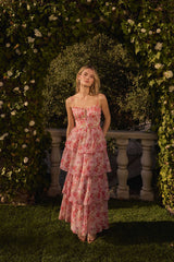 A young woman is wearing a long, tiered, pink floral dress with thin spaghetti straps. The dress features a fitted bodice with a square neckline and a smocked detail at the center. The skirt has multiple ruffled layers, each with a pink and white floral pattern. She is standing outdoors on a grassy area, framed by a lush green arch of climbing plants with small white flowers, and a stone balustrade is visible behind her.
