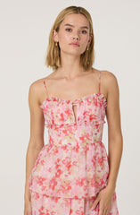 A woman is wearing a sleeveless, spaghetti strap top with a ruffled neckline and a small keyhole opening at the center. The top features a pink, red, and beige floral pattern. She is also wearing coordinating pants with the same floral print.