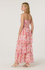 A woman is wearing a sleeveless, ankle-length dress with thin spaghetti straps. The dress features a pink and coral floral pattern with multiple tiered ruffles. She is also wearing white, open-toe high-heeled sandals.