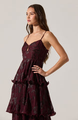 A woman is wearing a sleeveless, burgundy dress with thin spaghetti straps. The dress features a fitted bodice with a small keyhole opening at the center, and a pleated skirt with layered ruffles that cascade down in tiers. The fabric has a shiny, metallic finish with a black abstract pattern throughout.