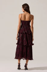 A woman is wearing a sleeveless, full-length dress with thin spaghetti straps. The dress is deep burgundy with a shiny, textured pattern, featuring multiple layered, ruffled tiers that add volume and movement. The fabric appears to be lightweight and semi-transparent with a delicate, slightly crinkled appearance. She pairs the dress with dark high-heeled shoes with ankle straps.