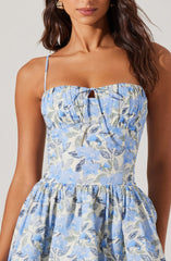 A woman is wearing a sleeveless, square-neck dress with thin spaghetti straps. The dress features a light blue floral print with shades of blue, green, and beige, and has a gathered bodice with a small keyhole and tie detail at the neckline. The skirt is full and gathered at the waist, creating a voluminous silhouette.