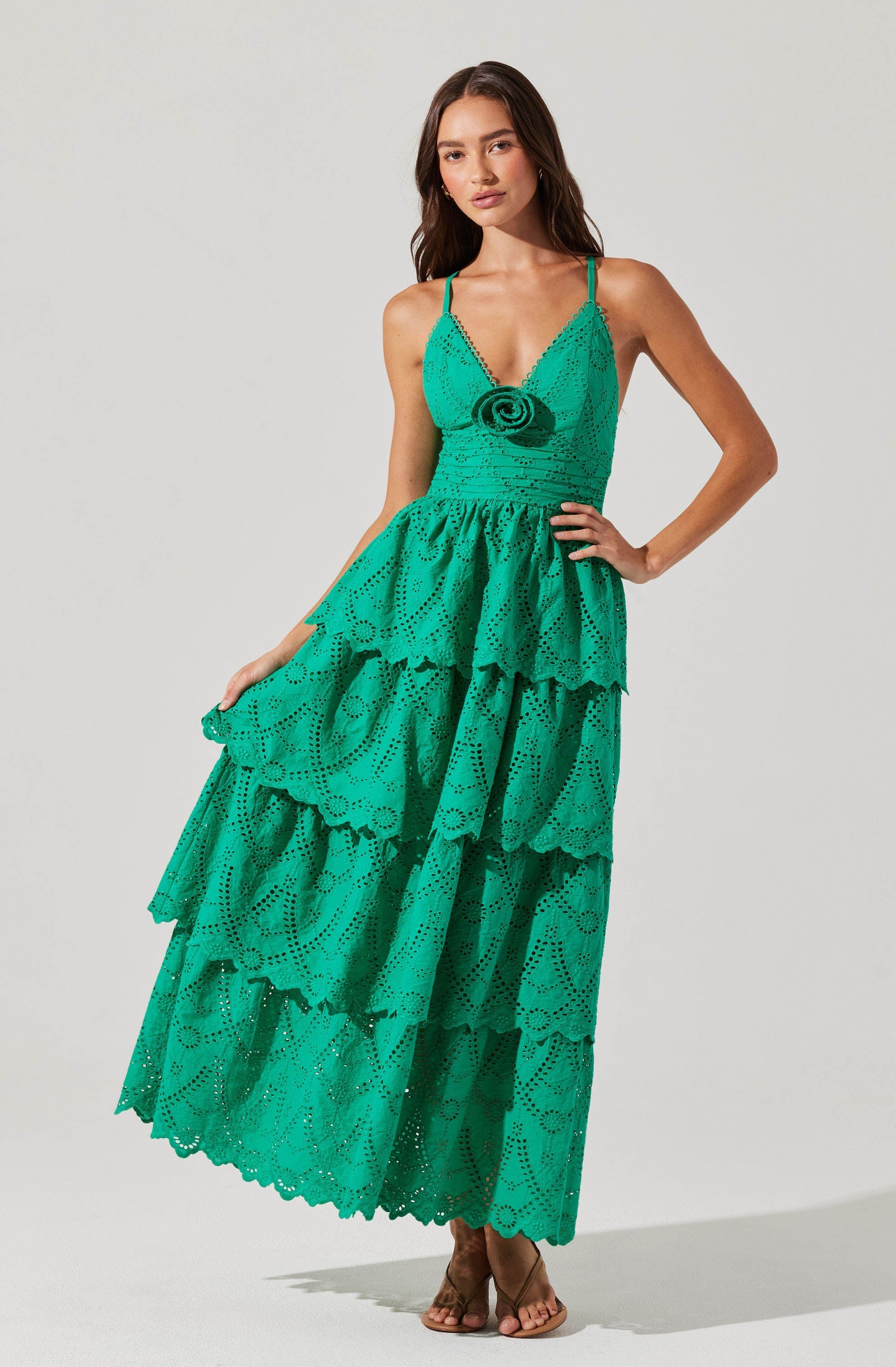 A woman is wearing a vibrant, turquoise, eyelet maxi dress with tiered ruffles. The dress features a fitted bodice with thin spaghetti straps, and a decorative rosette detail at the front. The skirt has multiple layers with scalloped edges, creating a flowing, tiered silhouette.