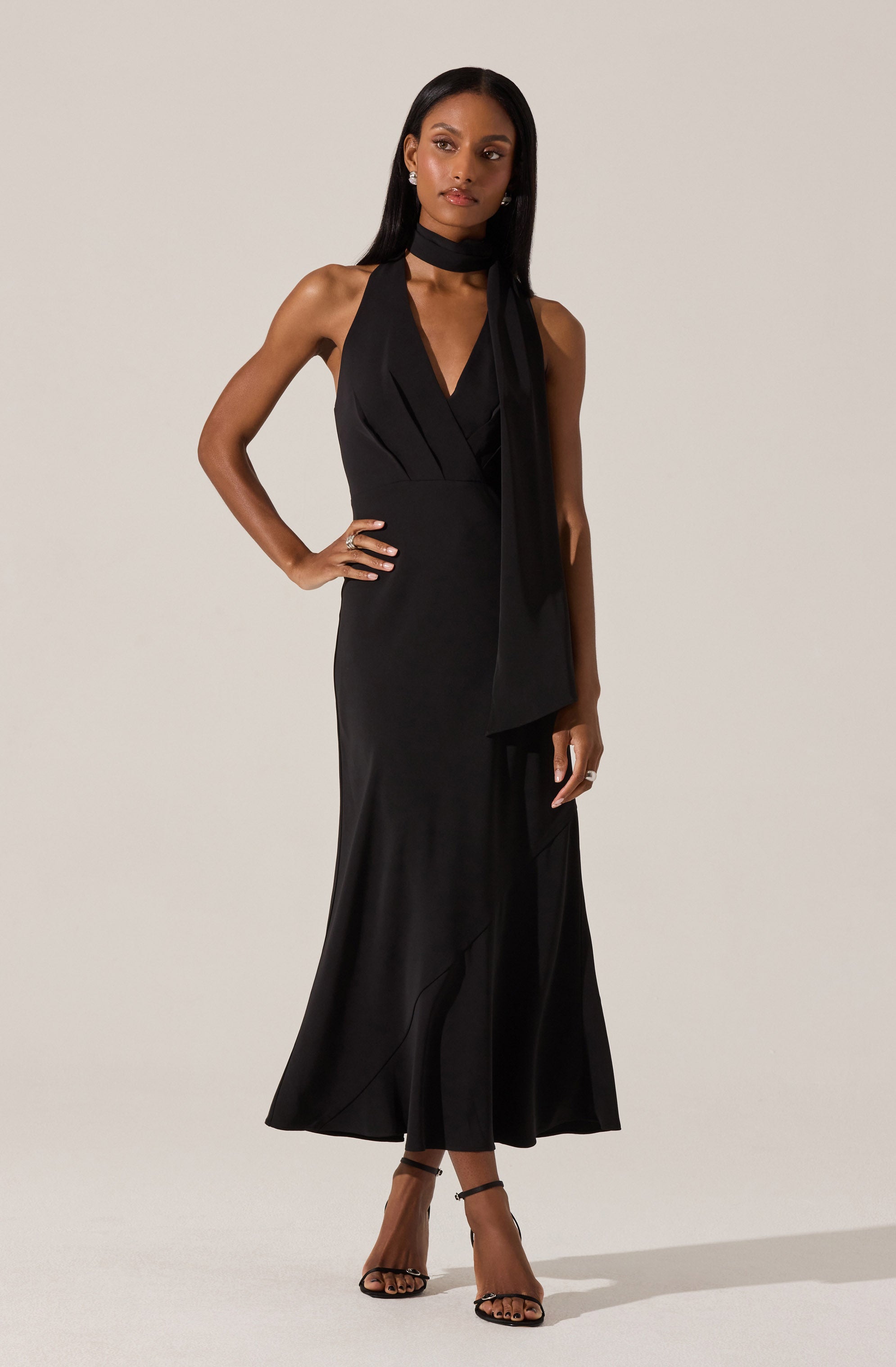 A woman is wearing a sleek, black sleeveless gown with a deep V neckline and a fitted waist. The dress features a long, flowing skirt that reaches the floor with subtle wave-like details. She has accessorized with a matching black choker and is wearing open-toed high heels with ankle straps.