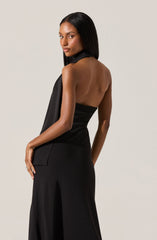 A woman is wearing a sleeveless, backless black dress with a high collar that drapes down one side. The dress is fitted at the waist and features a slit at the front of the skirt. The fabric looks smooth and shiny, giving an elegant appearance.