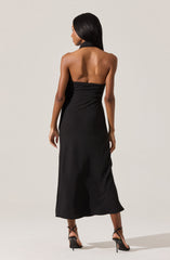 A woman is wearing a sleeveless black dress with a halter neck and an open back. The dress is fitted at the waist and flows down to a midi length. She is also wearing black high-heeled sandals with ankle straps.